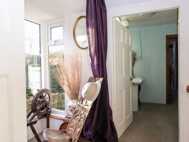 A corner with a wooden chair and spinning wheel near a window a vase with dried plants a round mirror on the wall and an open door showing a room with a sink at Castle Lodge Borrowdale