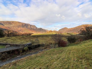 A grassy field with a gravel path and a wooden gate surrounded by hills and trees at Castle Lodge in Borrowdale