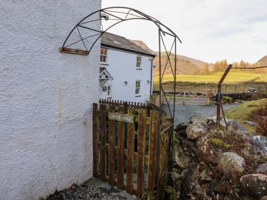 A wooden gate with a metal arch next to a white building and stone wall at Castle Lodge in Borrowdale