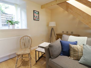 A sitting area with a wooden chair next to a window a sofa with cushions nested tables a tripod floor lamp and a wooden sideboard under a slanted ceiling at Y Cilgwyn in Trawsfynydd