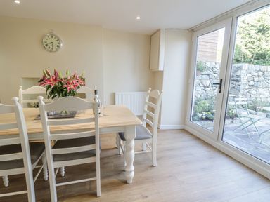 A dining area with a wooden table and white chairs and flowers on the table next to glass doors showing a stone patio at Y Cilgwyn in Trawsfynydd