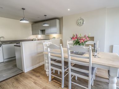 A dining area with a wooden table and white chairs next to a kitchen with hanging lights and cabinets at Y Cilgwyn in Trawsfynydd
