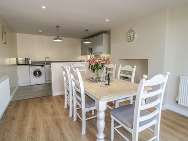 A dining area with a wooden table, white chairs, flowers, a bottle, and a kitchen with appliances in the background at Y Cilgwyn in Trawsfynydd