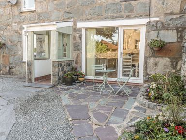 An outdoor patio area with stone flooring a small round table with two chairs potted plants and flowers and a stone building with a sliding glass door at Y Cilgwyn in Trawsfynydd