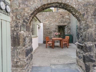 A stone archway leading to an outdoor patio with a wooden table and chairs and a vase with flowers at Y Cilgwyn in Trawsfynydd