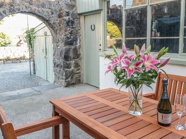 An outdoor area with a wooden table and chairs a vase with pink flowers a bottle of prosecco and empty glasses near stone arch and green painted doors at Y Cilgwyn in Trawsfynydd
