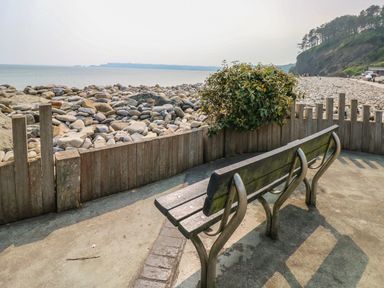 A wooden bench on a concrete path next to a wooden fence and rocky beach with sea and trees in the background at Pottery Cottage in Begelly near Kilgetty