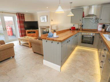 A kitchen with wooden countertops and grey cabinets next to a living area with armchairs and a TV at Pottery Cottage in Begelly near Kilgetty