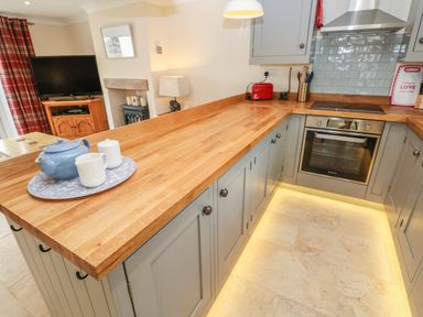 A kitchen with wooden countertops grey cabinets an oven a blue teapot on a tray and a living area with a wooden TV stand at Pottery Cottage Begelly near Kilgetty