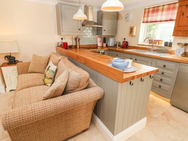 A kitchen with grey cabinets a wooden countertop a beige plaid sofa and a window with a checked blind at Pottery Cottage in Begelly near Kilgetty