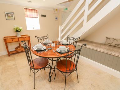 A dining area with a round wooden table set for four under stairs seating and a wooden side table with flowers at Pottery Cottage in Begelly near Kilgetty
