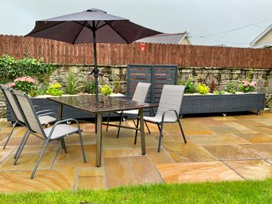 An outdoor patio area with a table and four chairs under a large umbrella and planters along a stone wall at Pottery Cottage in Begelly near Kilgetty