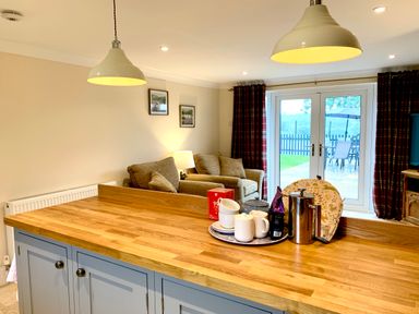 A kitchen island with cups and a teapot with a living area featuring armchairs and a lamp beside glass doors at Pottery Cottage in Begelly near Kilgetty