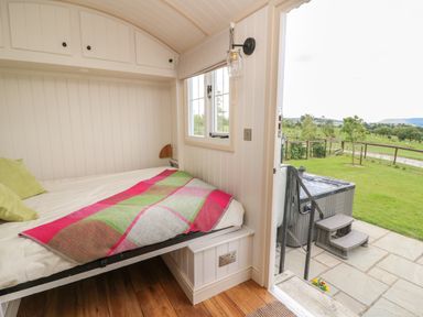 A small bedroom with a bed near a window and an open door showing a patio with a hot tub and a grassy field outside at Shepherd's View in Rhayader