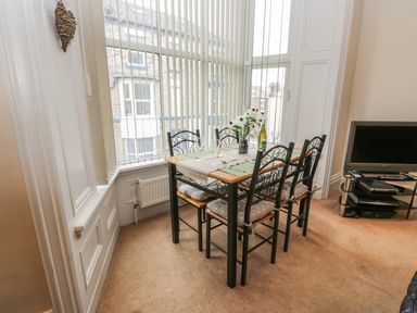A dining area with a table set for four by large windows with vertical blinds and a television on a stand at Flat 4 in Whitby