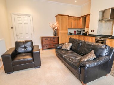 A living area with a black leather armchair and sofa with cushions next to a wooden kitchen and wooden chest of drawers at Flat 4 Whitby