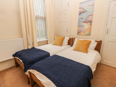 A bedroom with two single beds each with a blue blanket and yellow pillow a window with curtains and a painting on the wall at Flat 4 in Whitby