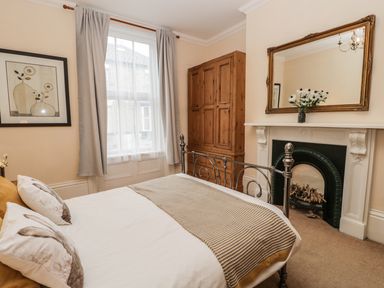 A bedroom with a bed pillow and blanket next to a window with curtains and a wooden wardrobe by a fireplace with a mirror above at Flat 4 in Whitby