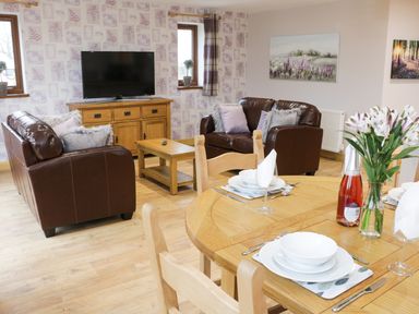A living room with brown leather sofas a wooden coffee table a wooden TV stand with a television and a dining table set with white dishes and flowers at Gardener's Lodge in Nantwich