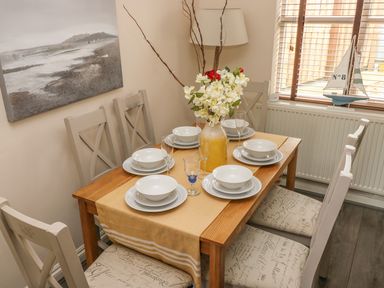 A dining table set for six with white plates bowls glasses and a flower vase in a room with six chairs a painting and a window with blinds at Sandy Bay House in Saundersfoot