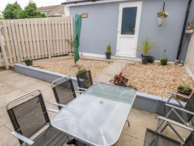 An outdoor patio with a glass table and four chairs near a building with a white door and pebbled garden area at Sandy Bay House in Saundersfoot