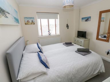 A bedroom with a double bed and white bedding towels on the bed a small TV on a dresser a window with blinds and wall art at Sandy Bay House in Saundersfoot