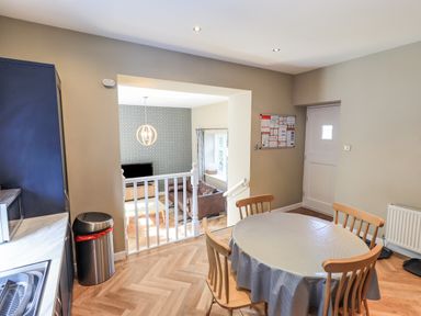 A dining area with a round table and four chairs next to a kitchen and a living room with a sofa TV and patterned wallpaper visible through an open wall at Loughrigg View in Ambleside