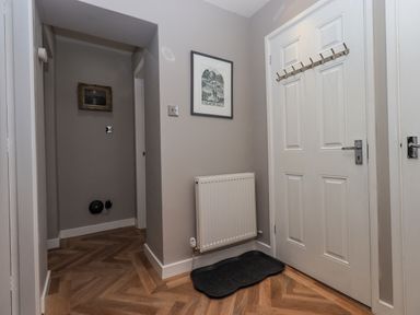 A hallway with a white door with hooks a radiator a black mat and framed pictures on the walls at Loughrigg View in Ambleside
