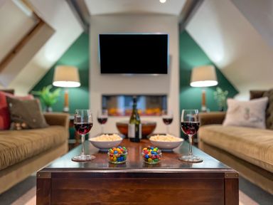 A living room with two sofas a wooden coffee table with wine glasses bowls of popcorn and jars of colorful candies a wall-mounted television and two lamps at Geltsdale East Wing in Wetheral