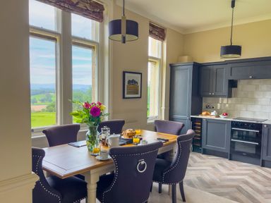 A kitchen and dining area with a wooden table set with breakfast items and windows showing countryside views at Geltsdale East Wing in Wetheral
