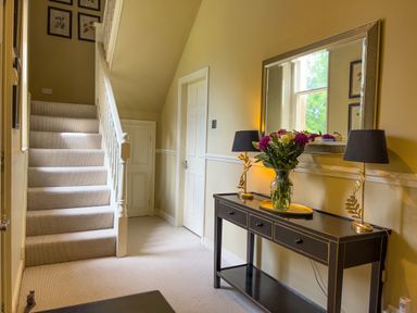 A hallway with carpeted stairs a black console table with lamps and a vase of flowers and a large wall mirror at Geltsdale East Wing in Wetheral