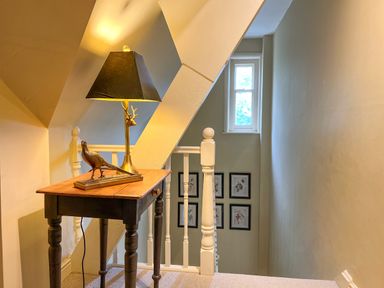 A small wooden table with a lamp and bird sculpture at the top of a staircase with framed pictures and a window in the background at Geltsdale East Wing in Wetheral