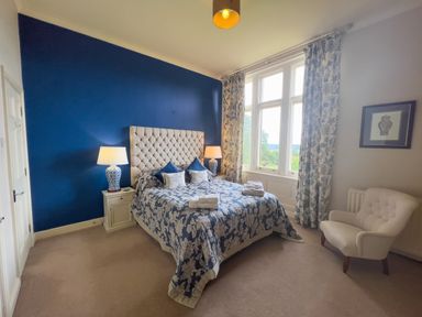A bedroom with a bed with blue floral bedding and beige headboard two bedside tables with lamps large window with floral curtains and an armchair at Geltsdale East Wing in Wetheral