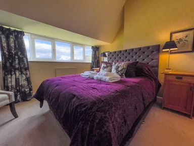 A bedroom with a large bed with a purple bedspread and towels on it two bedside tables with lamps and a window with floral curtains at Geltsdale East Wing in Wetheral