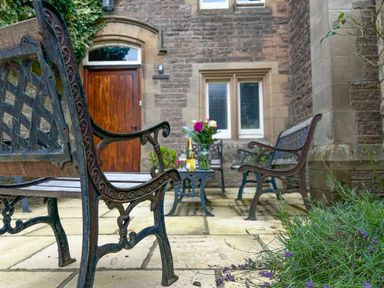 An outdoor patio with metal and wood benches a small table with flowers and drinks in front of a stone building at Geltsdale East Wing in Wetheral