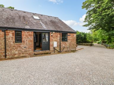 A stone house with black framed windows and a door opening to a gravel driveway surrounded by greenery at The Old Byre in Appleby-in-Westmorland