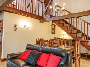 A living room with a black leather sofa and red cushions near a wooden dining table and staircase at The Old Byre in Appleby-in-Westmorland