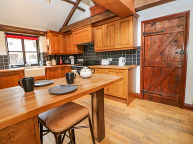 A kitchen with wooden cabinets a wooden island table two stools black mugs plates a teapot and a wooden door at The Old Byre in Appleby-in-Westmorland