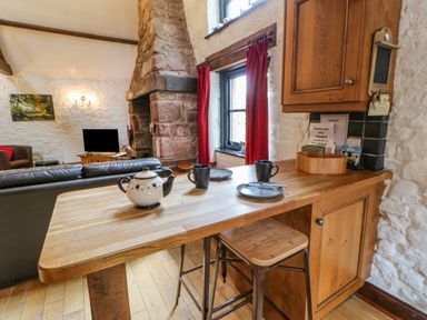 A kitchen counter with a teapot cups and plates next to a stool and a living area with a fireplace and television at The Old Byre in Appleby-in-Westmorland