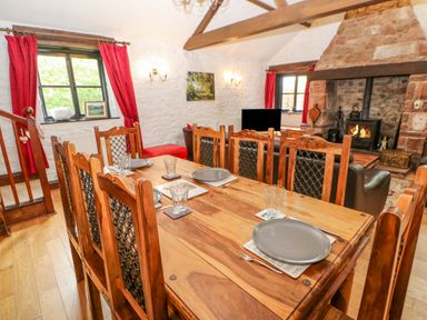 A dining room with a wooden table set with plates and glasses next to a seating area with a fireplace at The Old Byre in Appleby-in-Westmorland