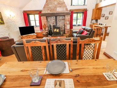 A dining table set with plates and glasses overlooking a living room with leather sofas a stone fireplace and red curtains at The Old Byre in Appleby-in-Westmorland