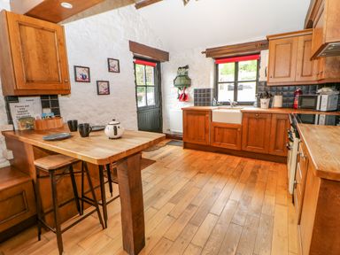 A kitchen with wooden cabinets a wooden floor a small wooden table with two stools and two windows at The Old Byre in Appleby-in-Westmorland