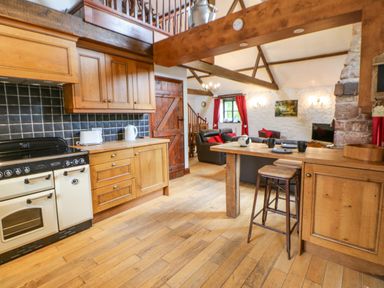 A kitchen with wooden cabinets a stove and a stool next to a wooden counter next to a living room with sofas and red curtains at The Old Byre in Appleby-in-Westmorland