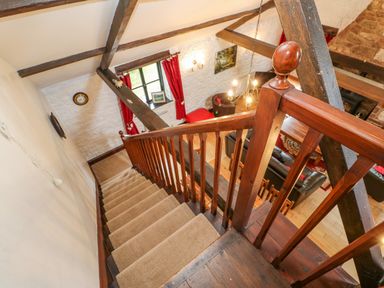 A staircase with wooden railing leading down to a living room with leather sofas wooden beams and red curtains at The Old Byre in Appleby-in-Westmorland