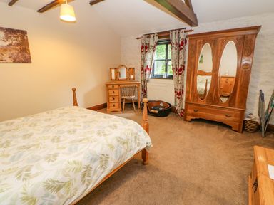 A bedroom with a wooden bed, a dressing table with a chair, a wardrobe with mirrors, and a pet bed near a window at The Old Byre in Appleby-in-Westmorland
