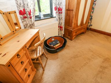 A room with a wooden desk and chair a window with floral curtains a wooden wardrobe and pet beds on the floor at The Old Byre in Appleby-in-Westmorland