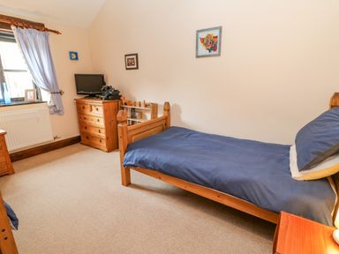 A bedroom with a single wooden bed with blue bedding a wooden chest of drawers a small bookshelf a window with checkered curtains and a beige carpet at The Old Byre in Appleby-in-Westmorland
