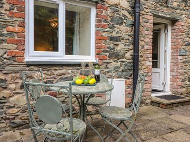 An outdoor patio area with a metal table and four chairs with a plate of fruit and a bottle of wine against a stone wall at Maple Cottage in Keswick