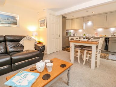 A living room with a black leather sofa a wooden coffee table with a teapot and book and a dining table set with four chairs in a kitchen area at Maple Cottage in Keswick