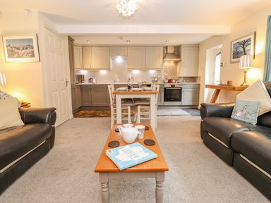 A living room with two black sofas and a wooden coffee table leading to a kitchen with grey cabinets and a dining table at Maple Cottage in Keswick